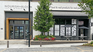 Nourish + Bloom's srong is painted white and has large windows. Trees are planted along the sidewalk in front of the store. Nourish + Bloom's srong is painted white and has large windows. Trees are planted along the sidewalk in front of the store.