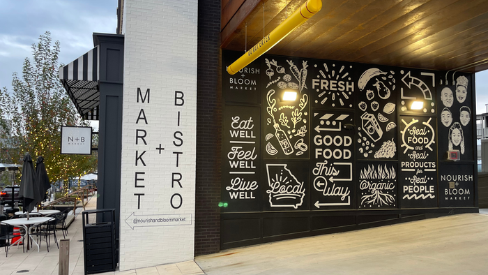 An outside wall of Nourish + Bloom with a black background is decorated with white paintings of produce, beverages and the Hemmings' family's faces. It also includes positive messages such as  An outside wall of Nourish + Bloom with a black background is decorated with white paintings of produce, beverages and the Hemmings' family's faces. It also includes positive messages such as