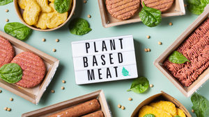 A sign that says "plant based meat" sits on a blue-green surface surrounded by yellow peas and a variety of packaged plant-based meats.  A sign that says "plant based meat" sits on a blue-green surface surrounded by yellow peas and a variety of packaged plant-based meats.