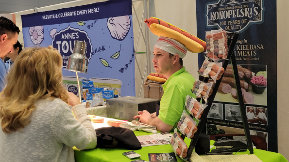 An exhibitor of natural meats wears a hat bearing a very large fabric hot dog in a bun.  An exhibitor of natural meats wears a hat bearing a very large fabric hot dog in a bun.
