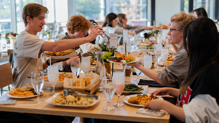 At a family-style meal in a cafeteria setting, students pass serving plates and bowls filled with pasta, chicken, green beans, breads and more. At a family-style meal in a cafeteria setting, students pass serving plates and bowls filled with pasta, chicken, green beans, breads and more.