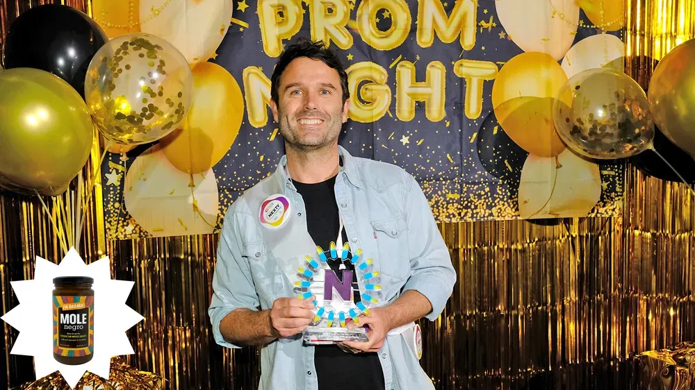 Azul Couzens, co-founder and CEO of ¡Ya Oaxaca!, poses with the brand's NEXTY Award trophy in front of a prom night banner, a gold backdrop and balloons.  Azul Couzens, co-founder and CEO of ¡Ya Oaxaca!, poses with the brand's NEXTY Award trophy in front of a prom night banner, a gold backdrop and balloons.