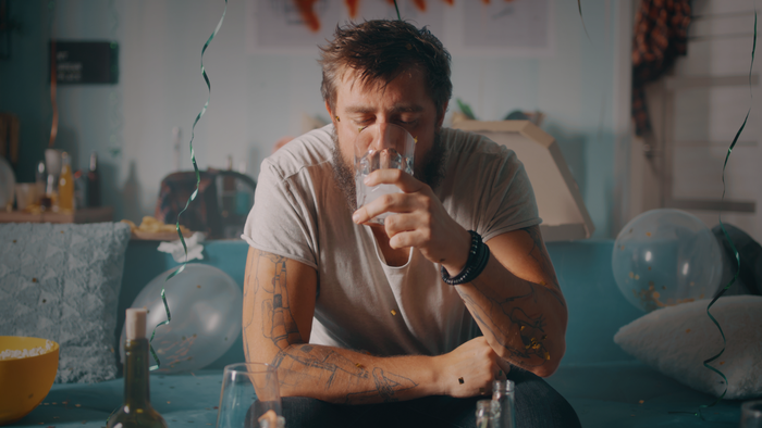 After a night of partying, a white man sits on his couch as sunlight peeks through an unseen window. He's nursing a drink with his eyes closed. Balloons and ribbons clutter the couch. After a night of partying, a white man sits on his couch as sunlight peeks through an unseen window. He's nursing a drink with his eyes closed. Balloons and ribbons clutter the couch.