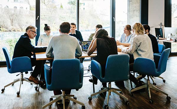 Nine people all sat around a meeting table talking, some using laptops