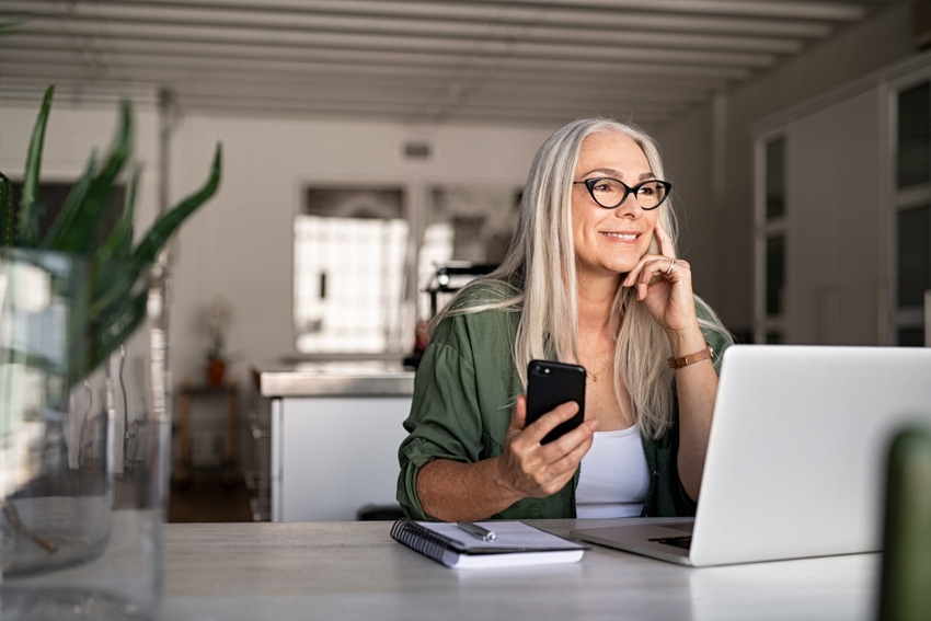 Happy senior woman holding smartphone and laptop daydreaming while looking away. Successful stylish old woman working at home while thinking about a