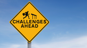 Photo illustration of a yellow road sign with the text "Challenges Ahead" printed below a silhouette of a businessman running toward a hurdle