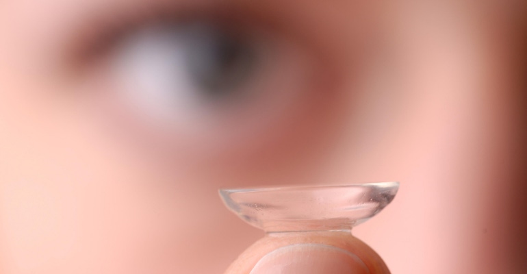 Image of a contact lens on a fingertip, with the wearer's eye blurred in the background.png