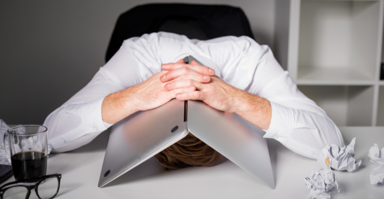 businessman with his head on his desk hiding underneath an open laptop.