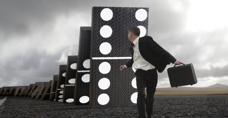 Business man running from giant falling dominoes.