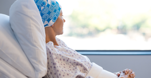 Female cancer patient in a hospital bed wearing a hospital gown and a blue head scarf looks out the window of her hospital