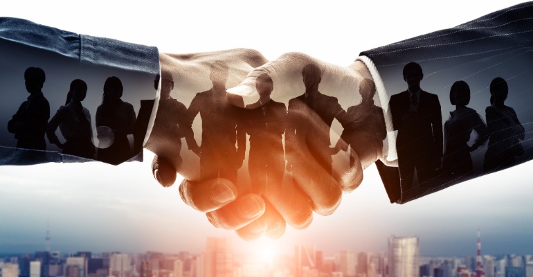 Business merger concept featuring a closeup of two business executives shaking hands with a team of employees overlaid.