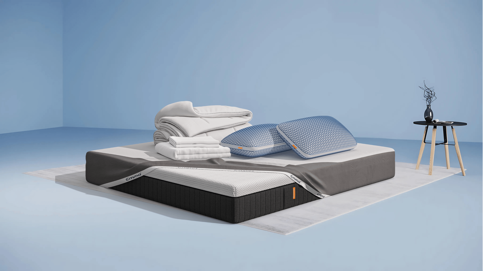 Ensemble de literie présenté dans une chambre moderne : matelas Hybride II ou Hybride Premium II avec base noire et surface blanche, partiellement recouvert d’un plaid gris foncé. Sur le matelas, deux oreillers Nuage Elite 40x70 cm bleus, une couette Nuage Tempérée blanche pliée, un protège-matelas, et une parure de lit en satin de coton blanc comprenant une housse de couette et des taies d’oreillers. Le tout est posé sur un tapis clair, avec une table d’appoint noire à pieds en bois et une décoration minimaliste.