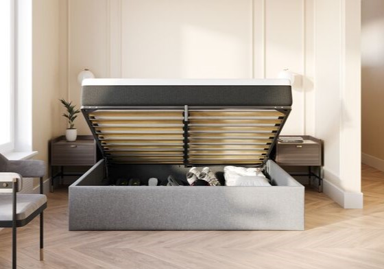 Lit coffre gris avec dessus à charnières ouvert, révélant un espace de rangement rempli de vêtements et de linge, dans une chambre moderne avec sol en bois et tables de chevet.