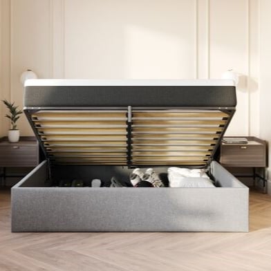 Lit coffre gris avec dessus à charnières ouvert, révélant un espace de rangement rempli de vêtements et de linge, dans une chambre moderne avec sol en bois et tables de chevet.