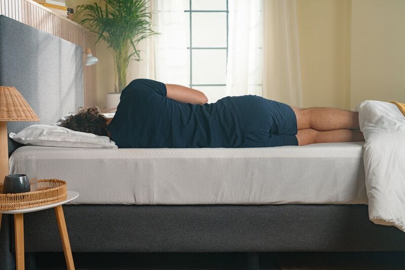 Person schläft seitlich auf einem Bett mit weißem Spannbettlaken in einem hellen Schlafzimmer.