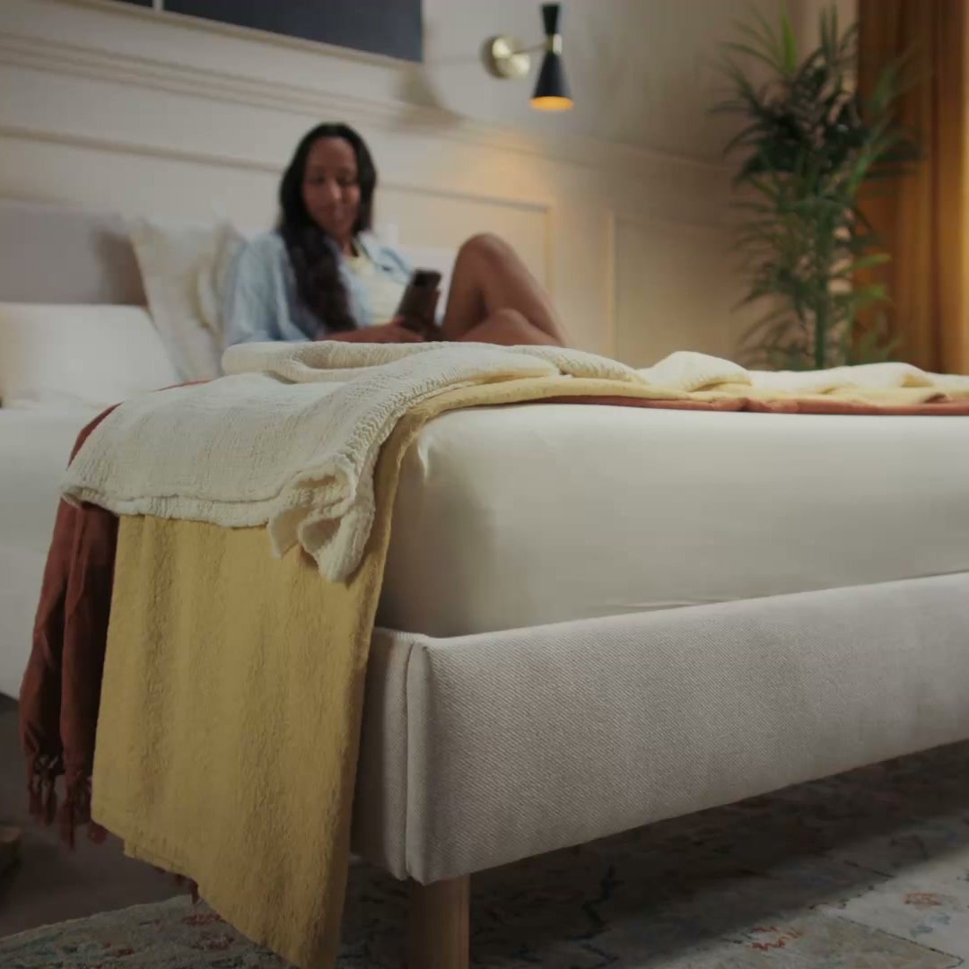 Woman relaxing on Emma light beige upholstered bed frame with natural wood legs in a warm bedroom.