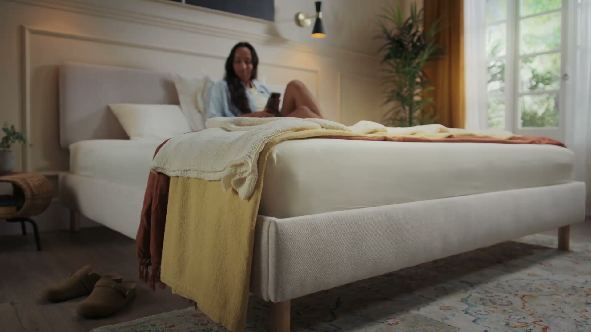 Woman relaxing on Emma light beige upholstered bed frame with natural wood legs in a warm bedroom.