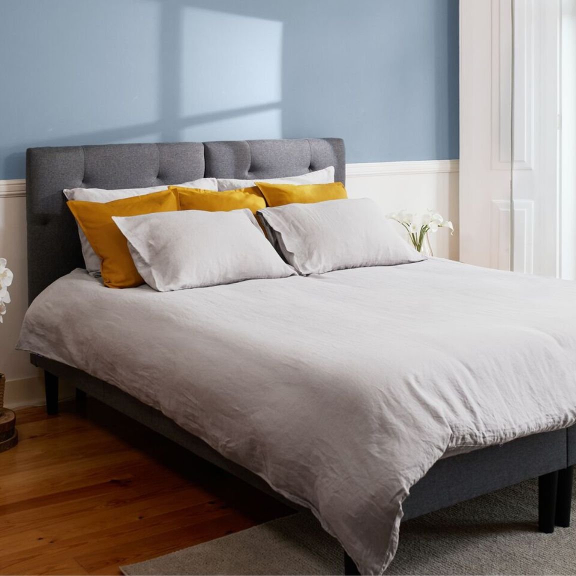Camera da letto ordinata con parete azzurra, letto con testiera grigia e decorazioni naturali.