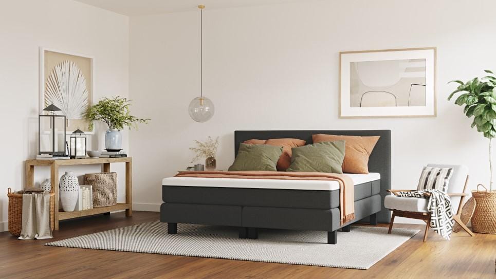 Stijlvolle slaapkamer met donkergrijs bedframe, witte matras en kussens in groen- en bruintinten; houten console met decoraties, witte fauteuil en grote groene plant bij witgeschilderde muur.