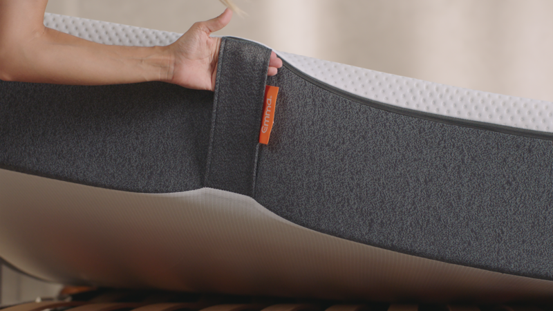 Close-up van een hand die de rand van een matras aanraakt, waarbij de grijze bekleding met textuur en witte bovenlaag zichtbaar zijn. Een oranje label met het merk 'Emma'. De matras ligt op een houten lattenbodem