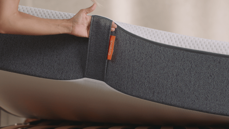 Close-up van een hand die de rand van een matras aanraakt, waarbij de grijze bekleding met textuur en witte bovenlaag zichtbaar zijn. Een oranje label met het merk 'Emma'. De matras ligt op een houten lattenbodem