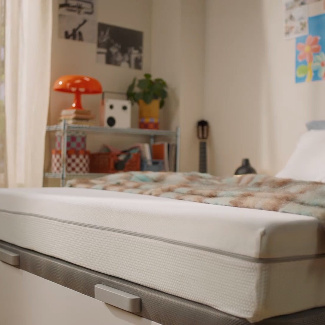 Person sitting on a bed with a white mattress in a bright bedroom.