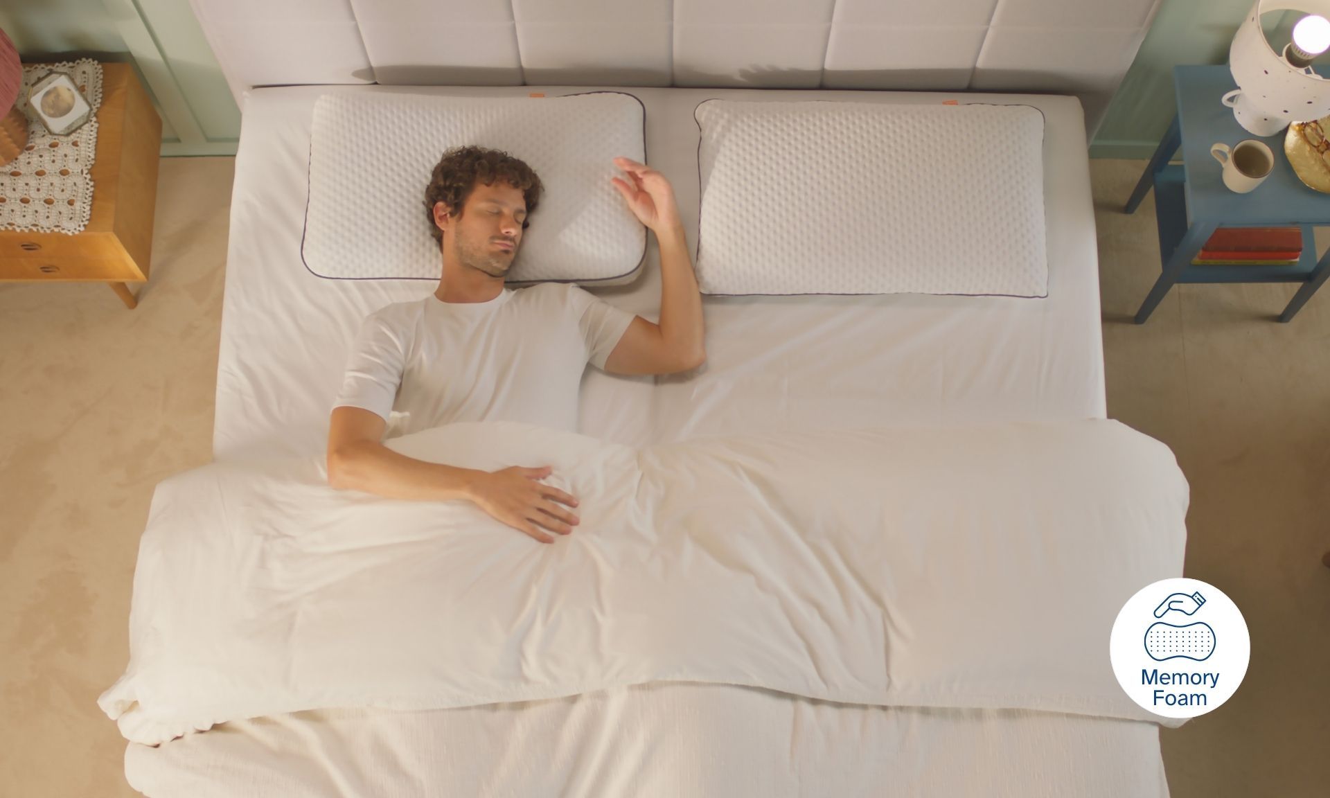 Persona sdraiata su letto con due cuscini, tavolini ai lati e icona 'Memory Foam' in basso a destra