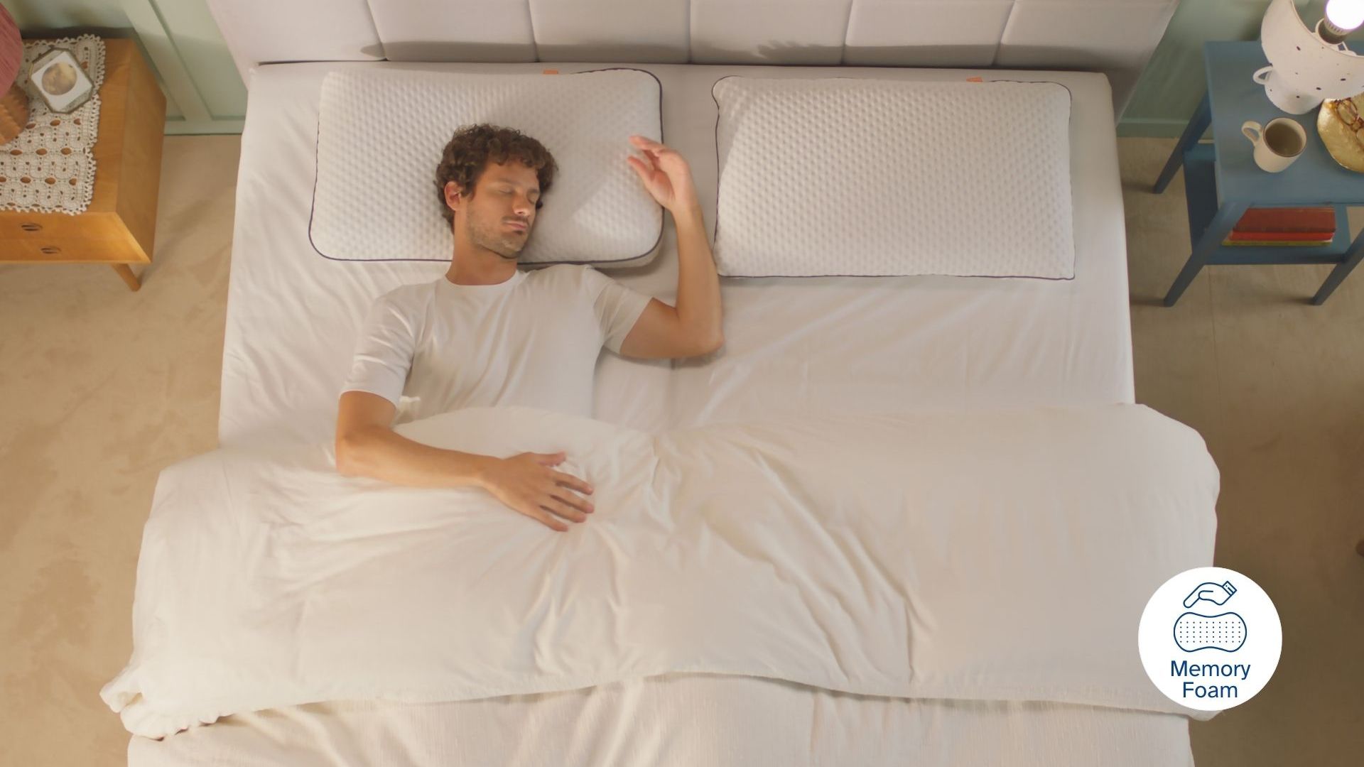 Persona sdraiata su letto con due cuscini, tavolini ai lati e icona 'Memory Foam' in basso a destra