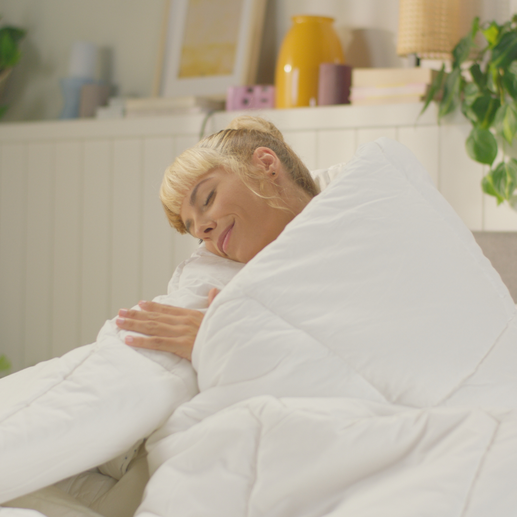 Personne assise dans un lit, enveloppée dans une grande couette blanche et confortable. À l'arrière-plan, une étagère avec des livres rouges, des objets décoratifs et des plantes vertes.