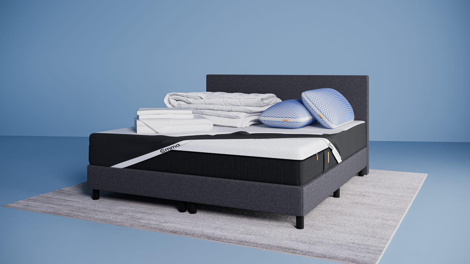 Un lit moderne avec un cadre et une tête de lit gris foncé, surmonté de deux oreillers bleus empilés, d'une literie blanche pliée et d'une couverture grise, se trouve sur un tapis clair dans une pièce bleue minimale, à côté d'une petite table d'appoint ronde.