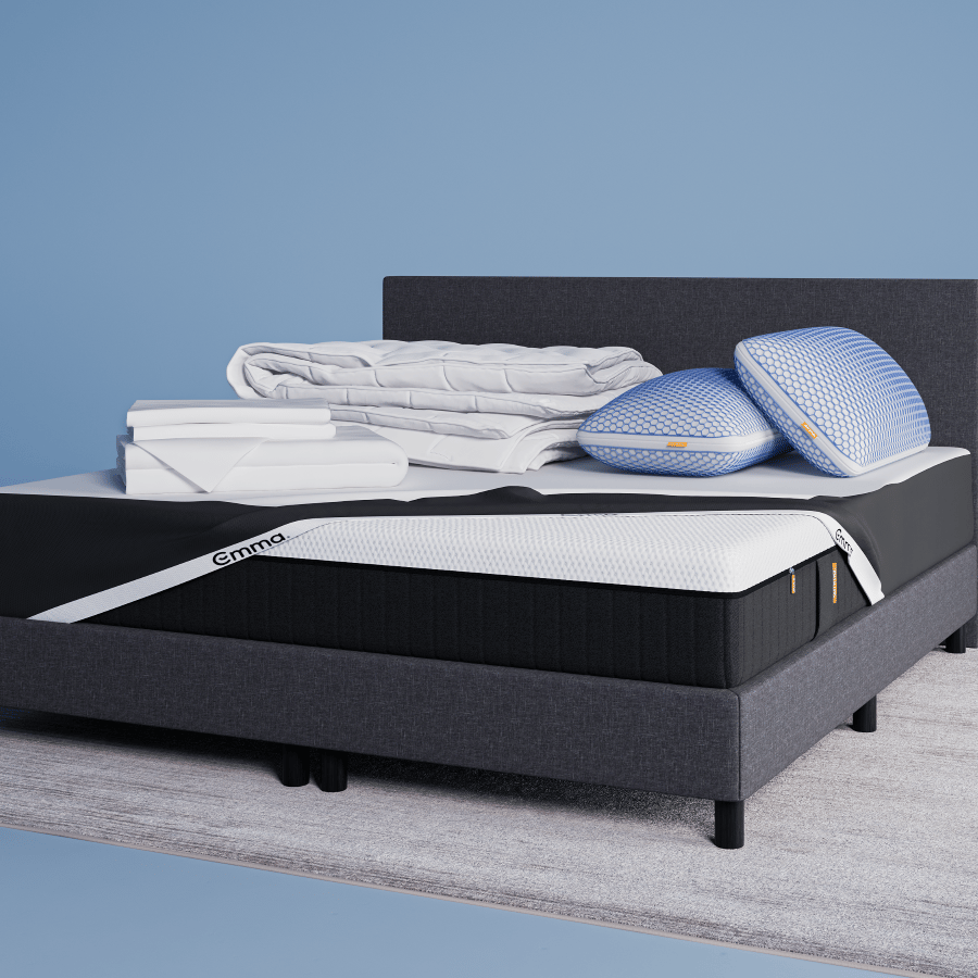 Un lit moderne avec un cadre et une tête de lit gris foncé, surmonté de deux oreillers bleus empilés, d'une literie blanche pliée et d'une couverture grise, se trouve sur un tapis clair dans une pièce bleue minimale, à côté d'une petite table d'appoint ronde.