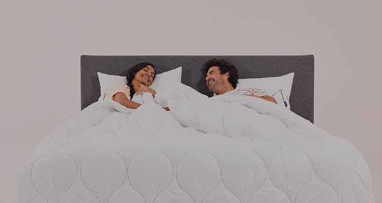 A couple lying in bed under a white duvet, smiling and looking at each other.