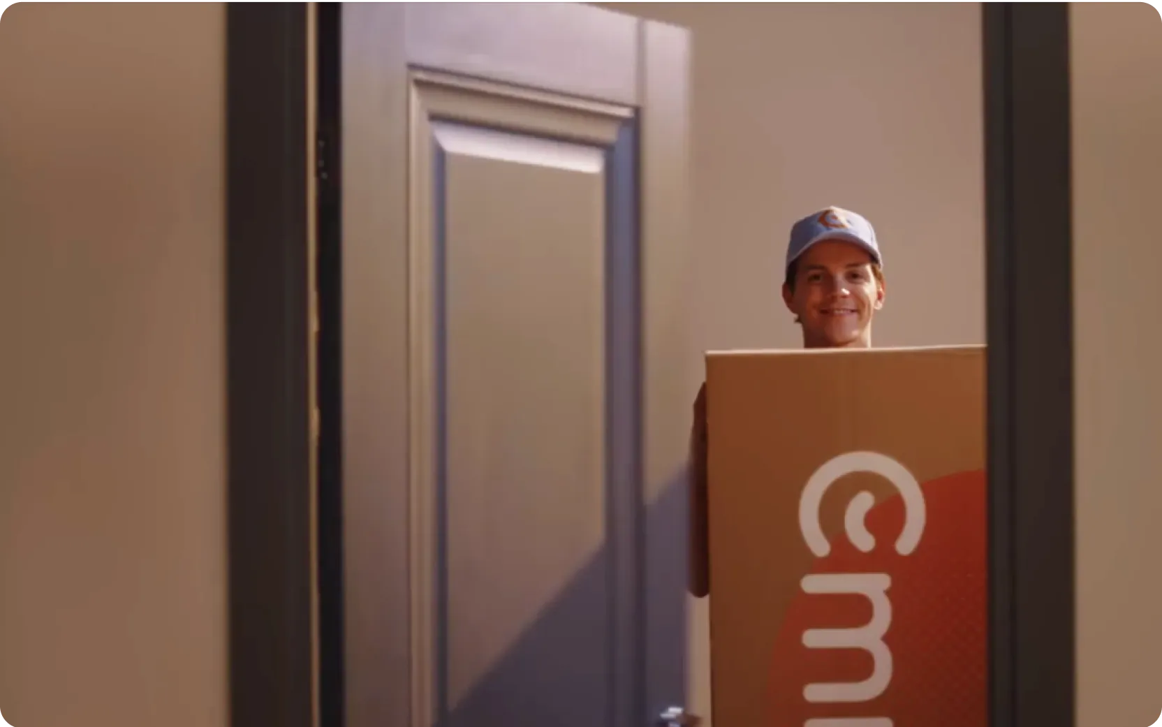Persoon staat glimlachend in een deuropening met een grote kartonnen doos met zichtbaar Emma-logo. De setting oogt als een levering of verhuismoment in een huiselijke omgeving.