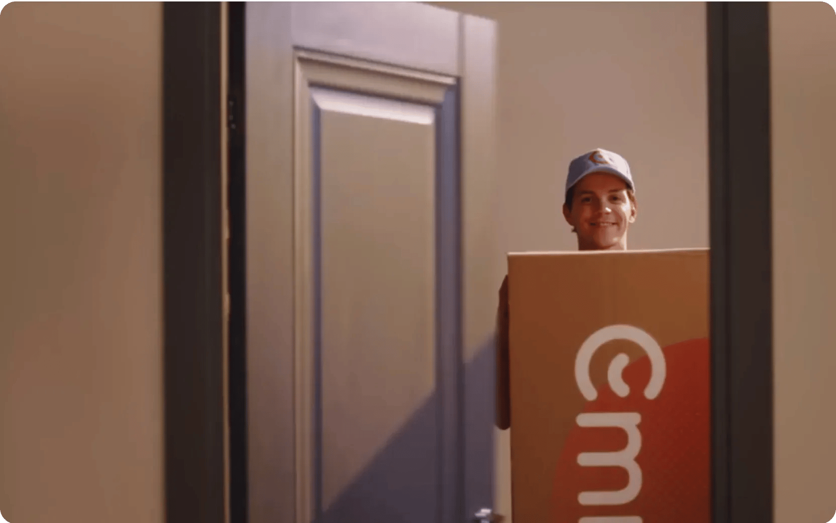 Persoon staat glimlachend in een deuropening met een grote kartonnen doos met zichtbaar Emma-logo. De setting oogt als een levering of verhuismoment in een huiselijke omgeving.