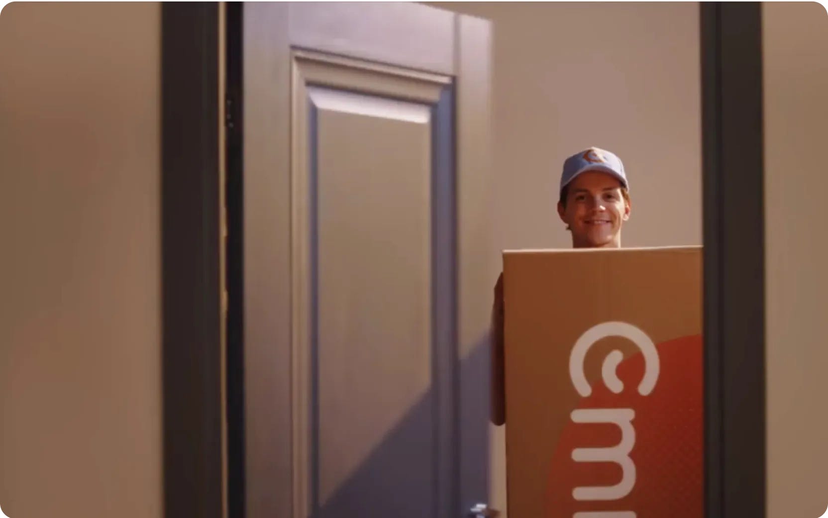 Persoon staat glimlachend in een deuropening met een grote kartonnen doos met zichtbaar Emma-logo. De setting oogt als een levering of verhuismoment in een huiselijke omgeving.