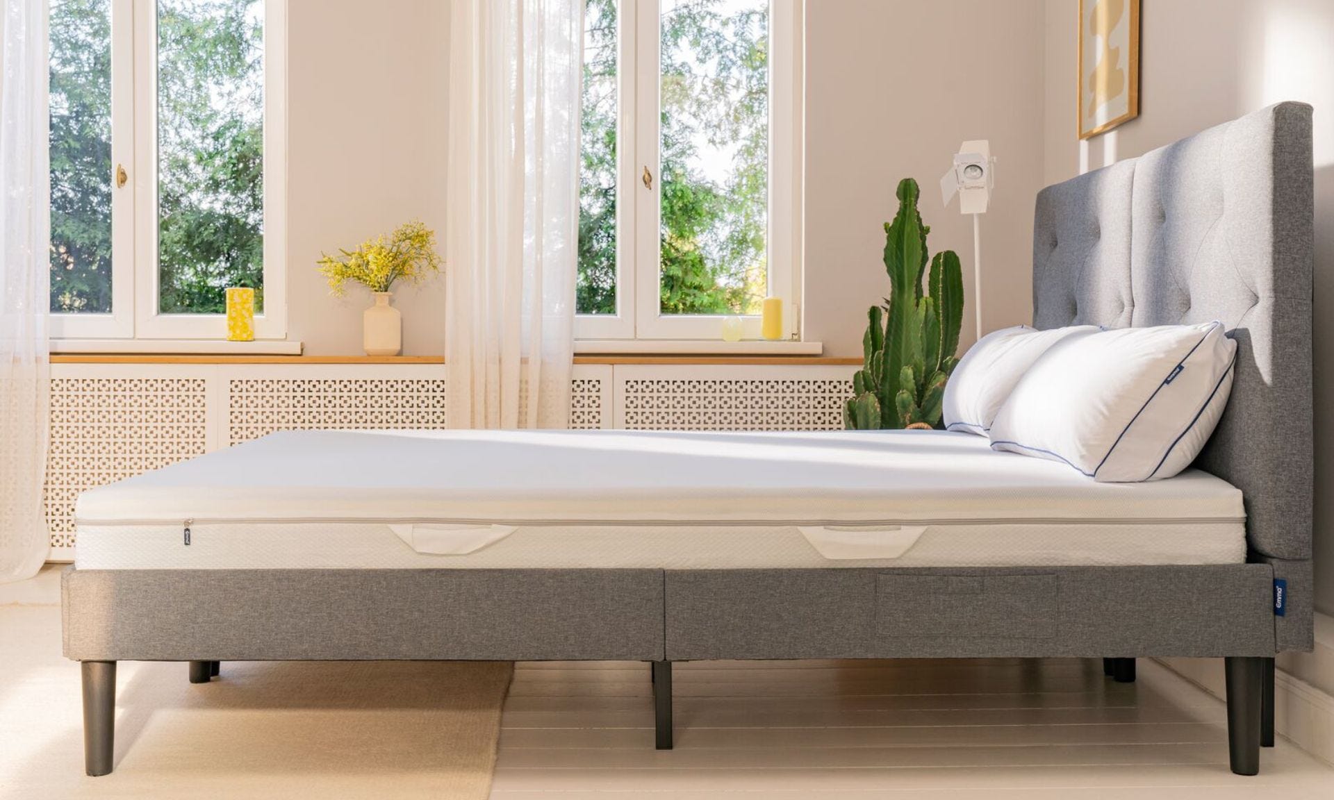 Letto con testiera grigia in stanza luminosa, decorato con fiori e piante.