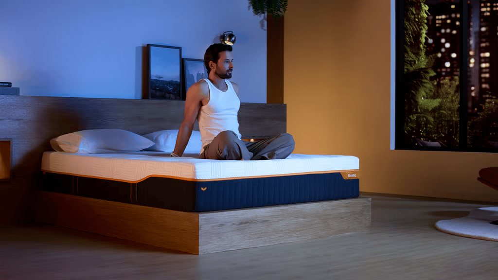 Man zittend op een Emma Performance Matras matras op een houten bedframe, in een sfeervolle slaapkamer met stadslichten op de achtergrond.