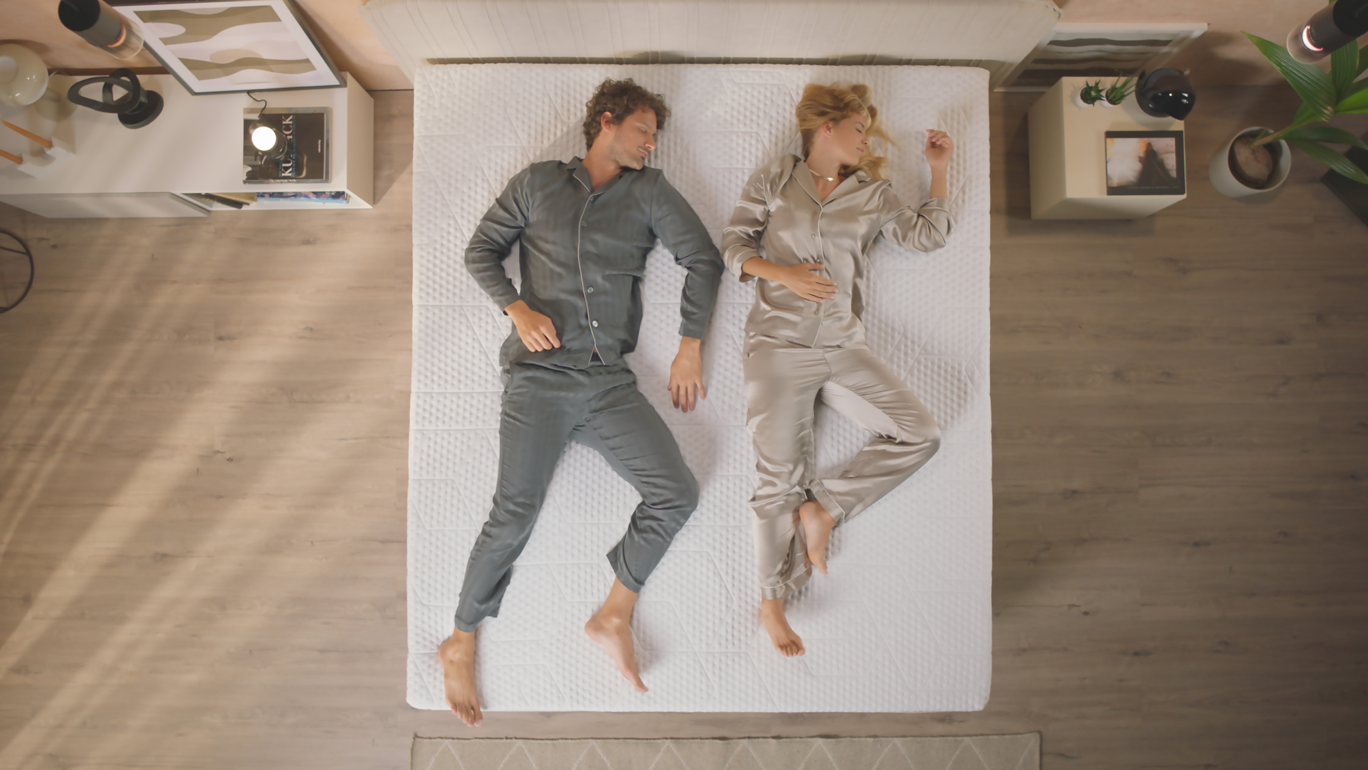 Due persone in pigiama sdraiate su un letto con topper bianco, comodini ai lati e tappeto ai piedi del letto