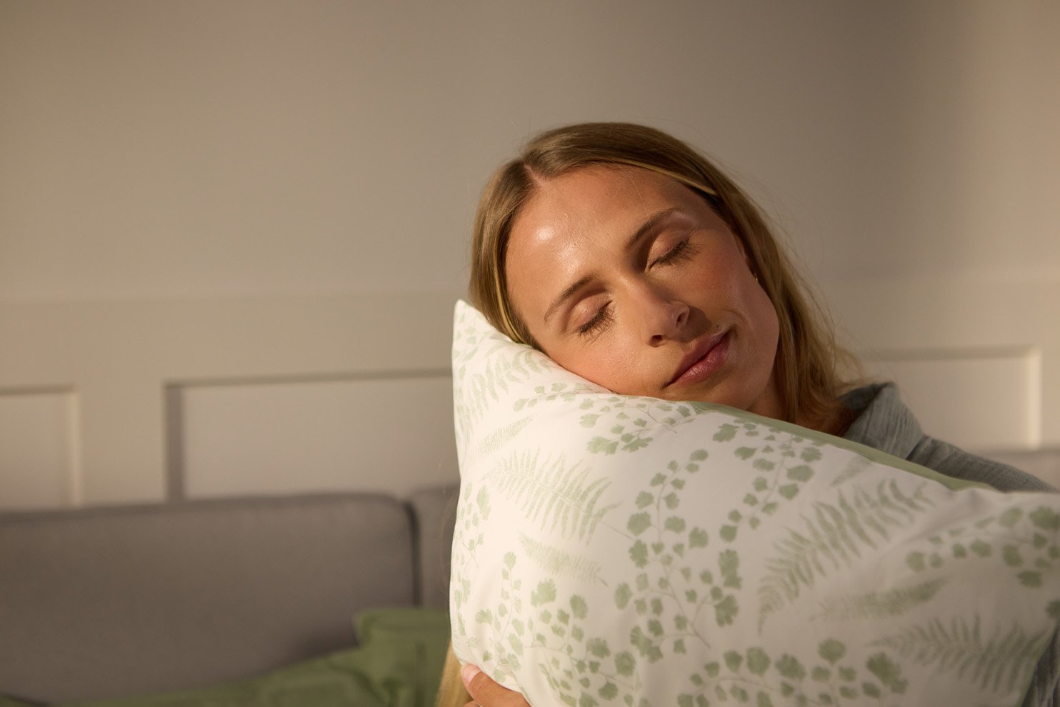Persoon houdt een kussen vast met lichtgroen botanisch patroon in een lichte slaapkamer.