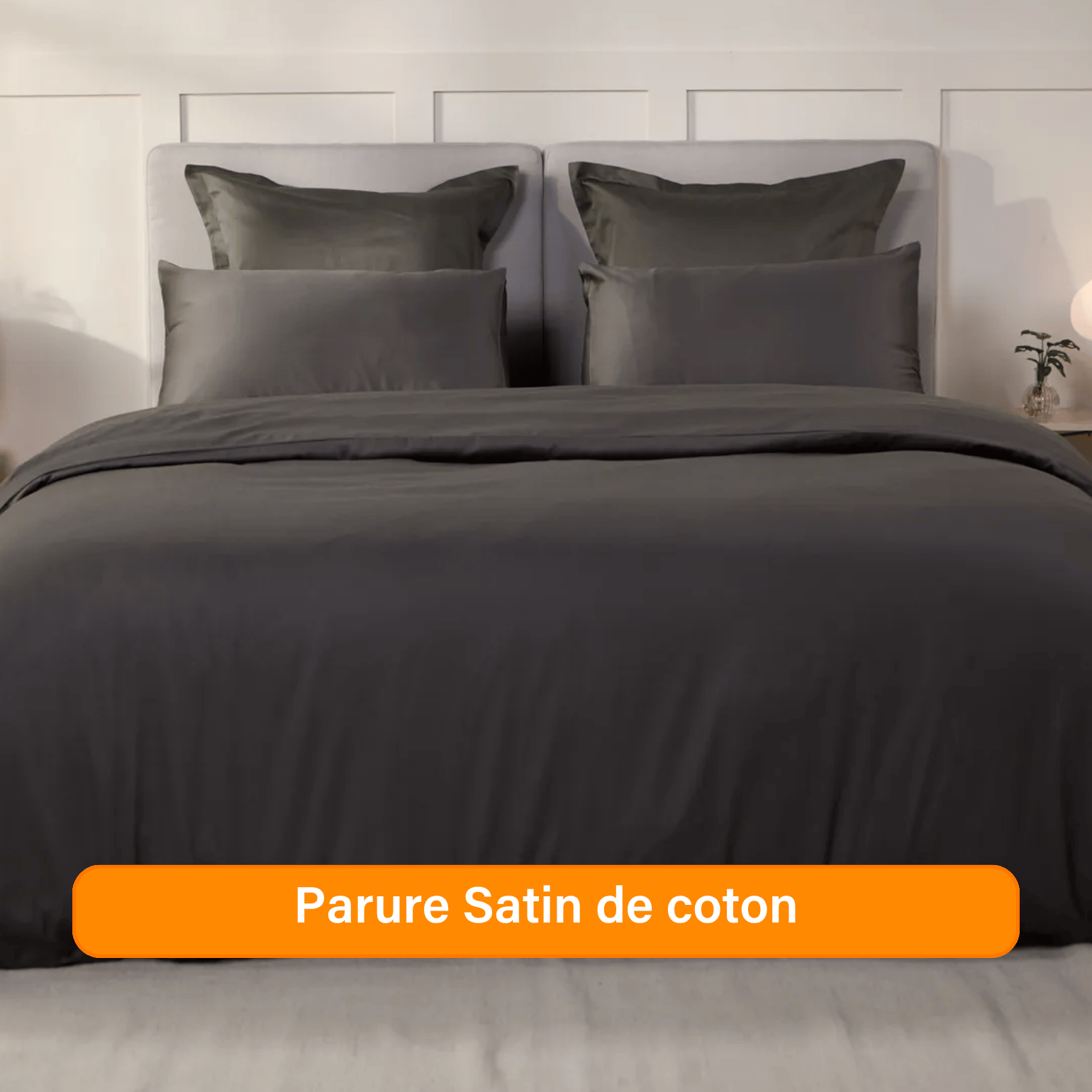 Chambre élégante avec lit soigneusement préparé, habillé d’une parure de lit 100 % satin de coton de couleur Gris Foncé. Plusieurs oreillers sont disposés contre un mur à panneaux. De chaque côté du lit, des tables de chevet avec lampe et objets décoratifs. Un banc rose est placé au pied du lit, assorti à un tabouret près de la table de chevet. Le sol en bois clair et les murs neutres créent une ambiance douce et raffinée.