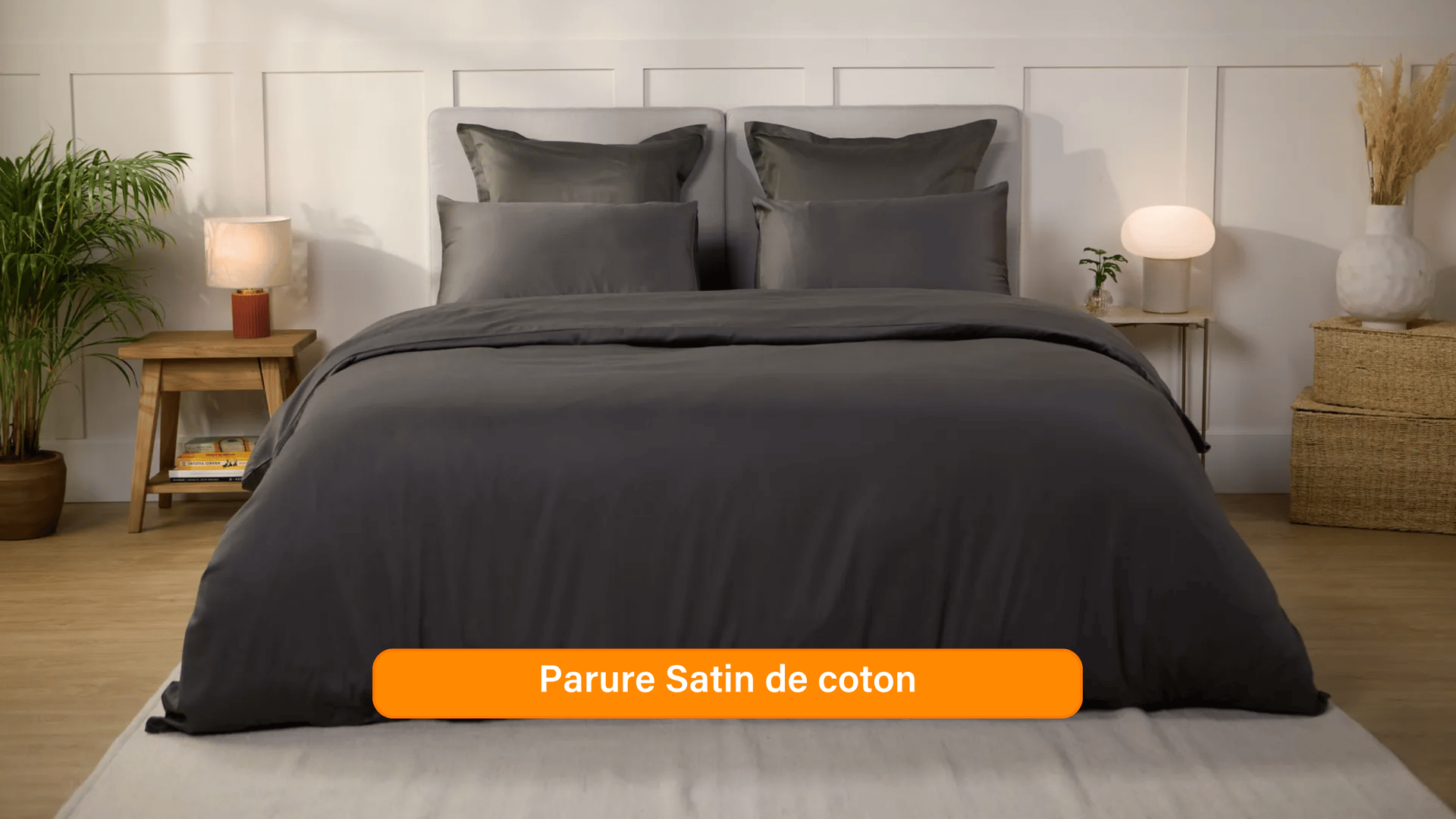 Chambre élégante avec lit soigneusement préparé, habillé d’une parure de lit 100 % satin de coton de couleur Gris Foncé. Plusieurs oreillers sont disposés contre un mur à panneaux. De chaque côté du lit, des tables de chevet avec lampe et objets décoratifs. Un banc rose est placé au pied du lit, assorti à un tabouret près de la table de chevet. Le sol en bois clair et les murs neutres créent une ambiance douce et raffinée.