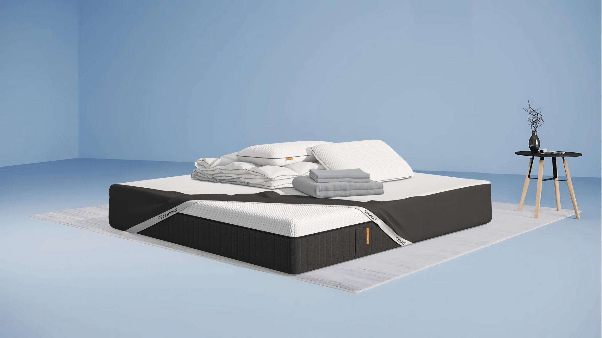 Matelas avec protège-matelas, deux oreillers, un drap plié, une couette et une housse de couette. Fond beige avec une forme orange abstraite.