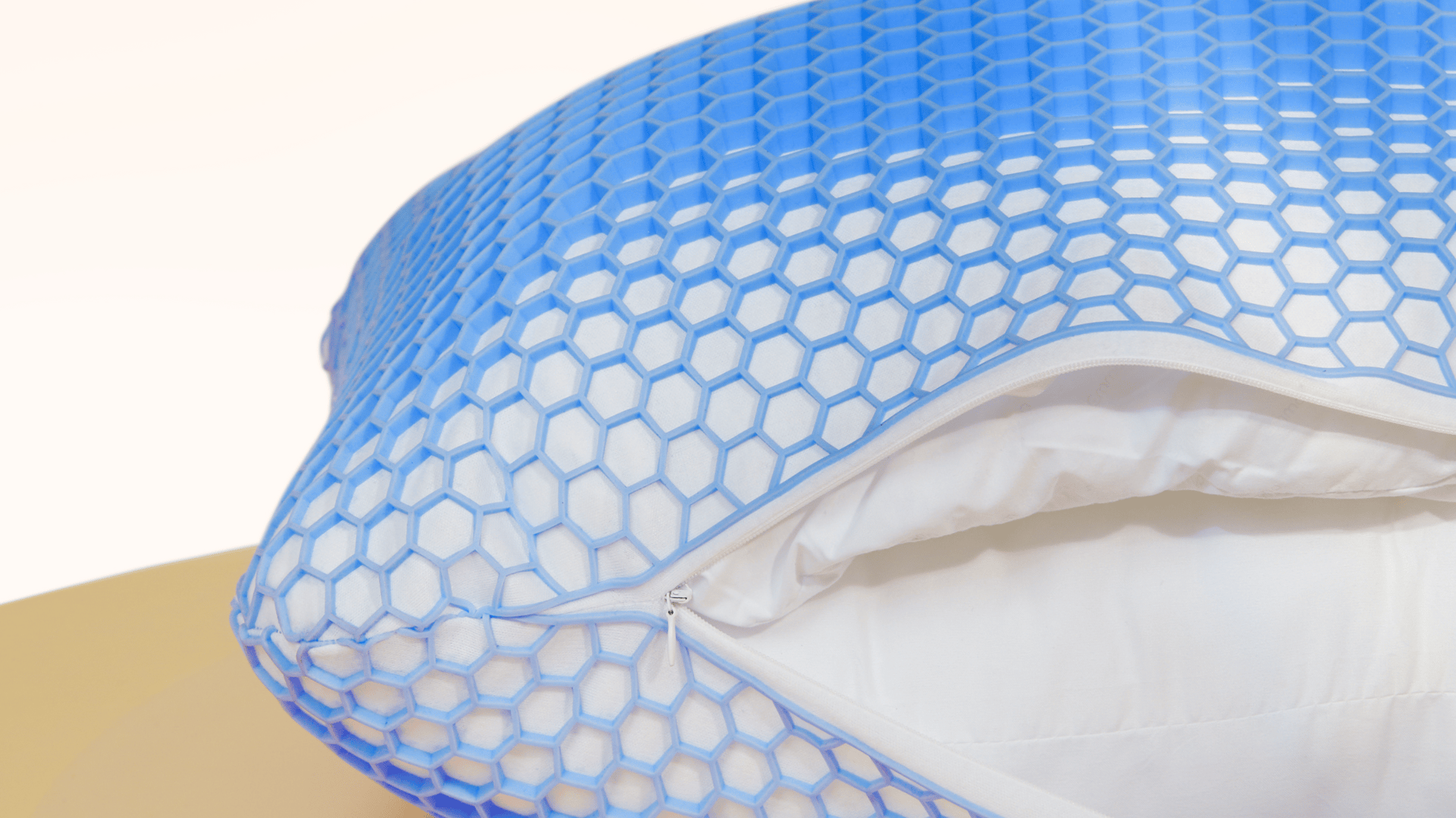 Gros plan sur un oreiller blanc dont la housse de gel hexagonale bleue est partiellement dézippée, montrant le tissu intérieur de l'oreiller. La housse de gel présente un motif en nid d'abeille et l'arrière-plan est légèrement éclairé.