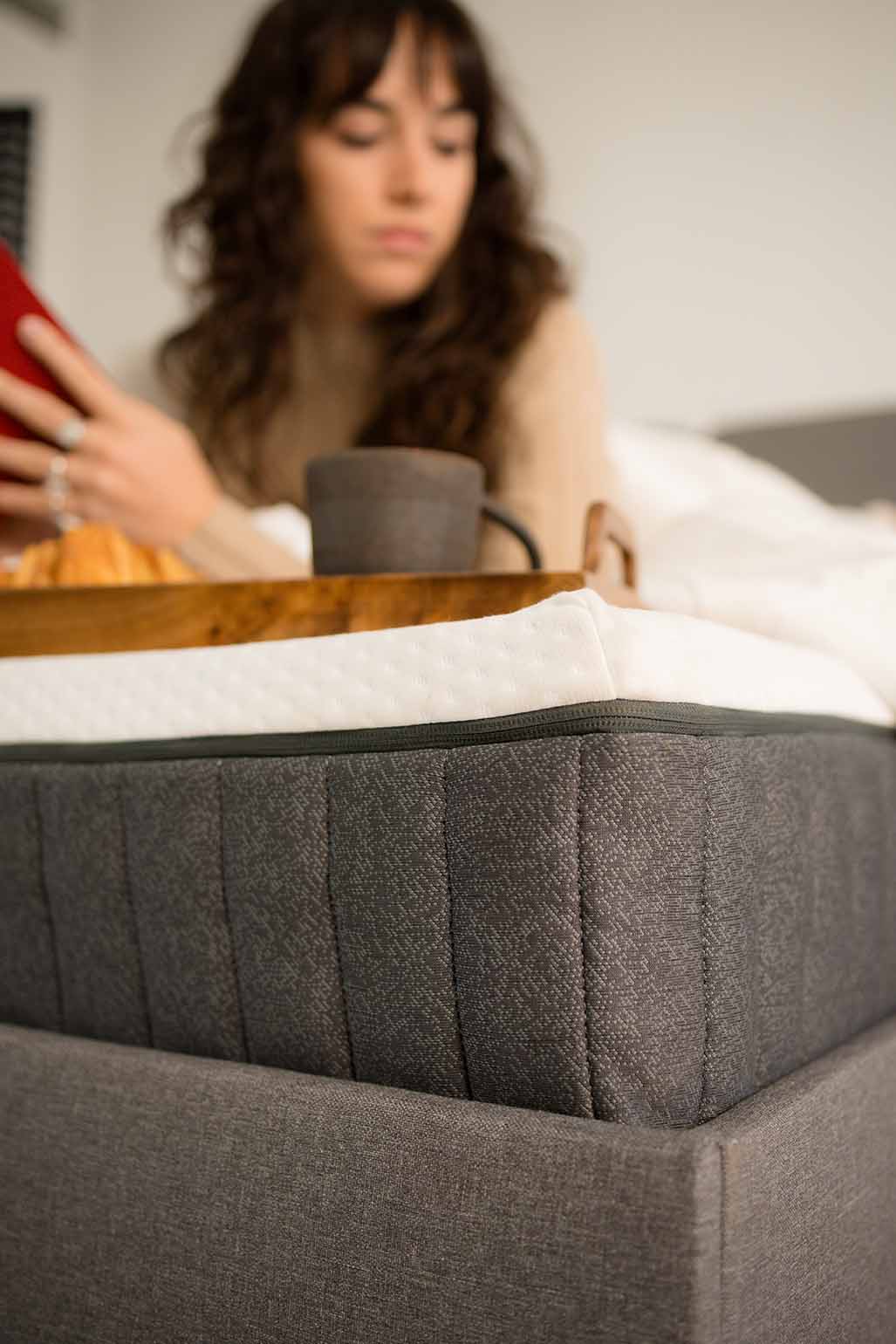 Letto con materasso grigio, vassoio in legno con tazza; persona sfocata sullo sfondo
