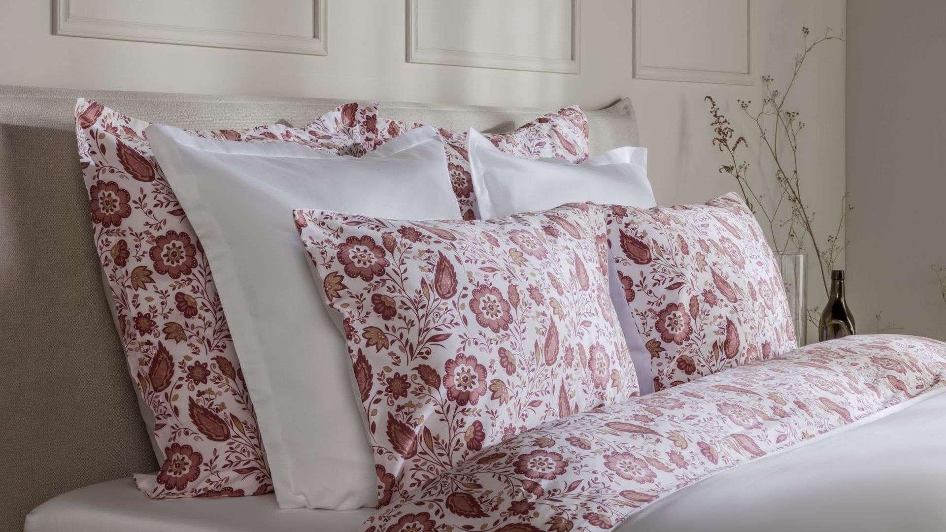 Letto ordinato con biancheria bianca e floreale nei toni del rosso e rosa, decorazioni naturali sullo sfondo.