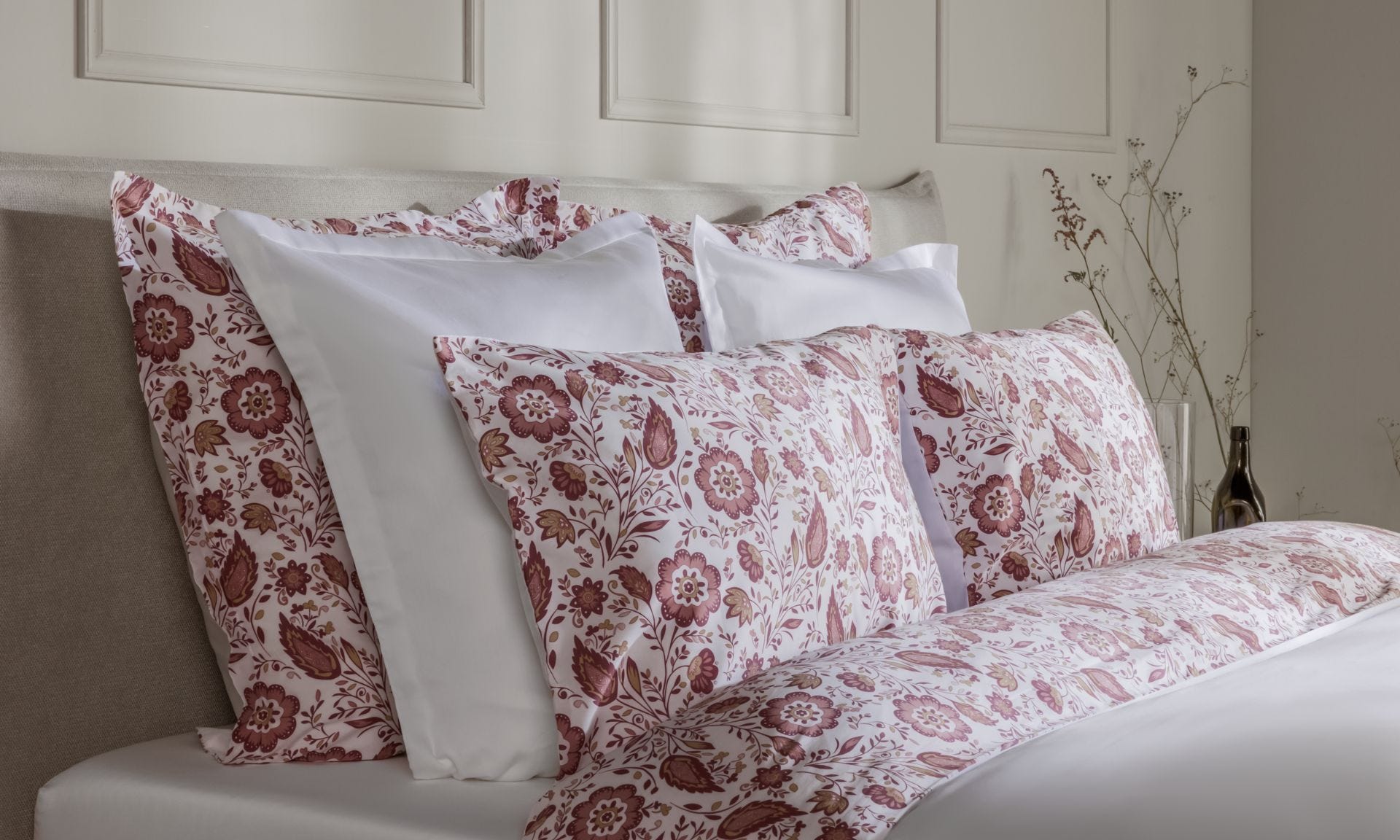 Letto ordinato con biancheria bianca e floreale nei toni del rosso e rosa, decorazioni naturali sullo sfondo.