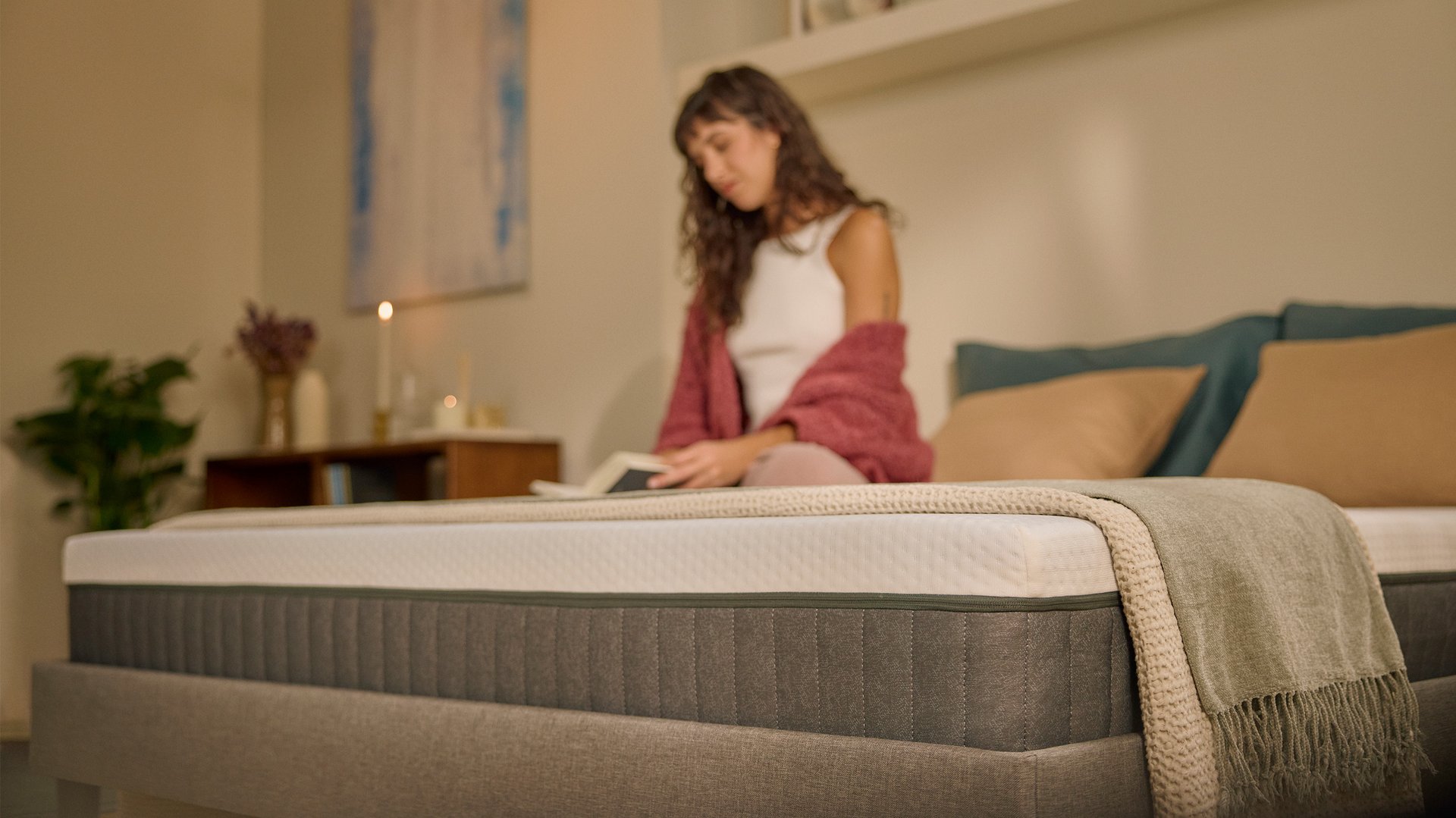 Une femme assise sur un matelas Emma Original Lite dans une chambre à l'éclairage chaleureux, avec un gros plan sur le matelas au premier plan.