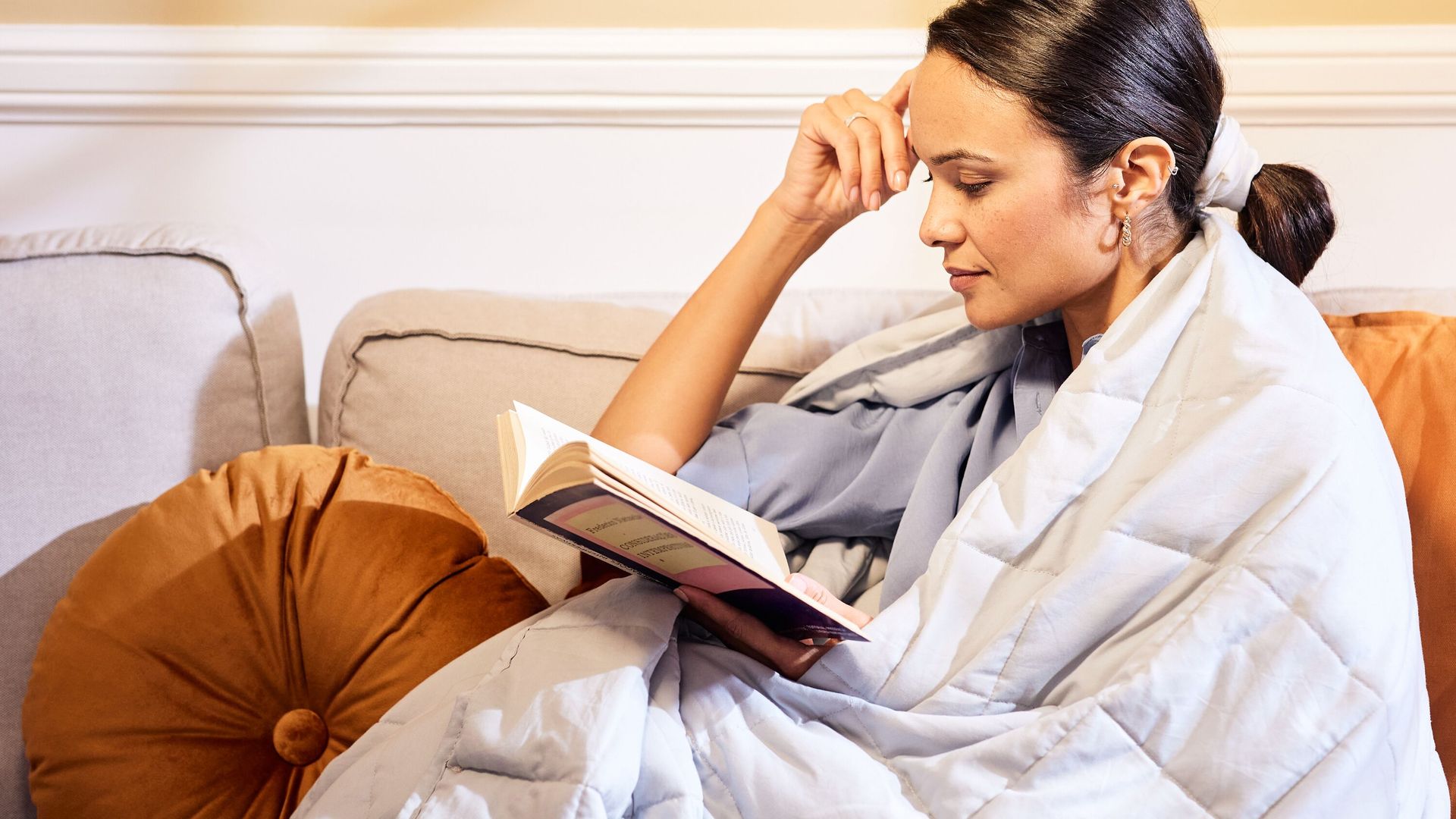 Persoon zit op bank onder lichtgrijze Emma verzwaringsdeken met oranje kussen naast zich—gezellige setting met boek en ontspannen sfeer.