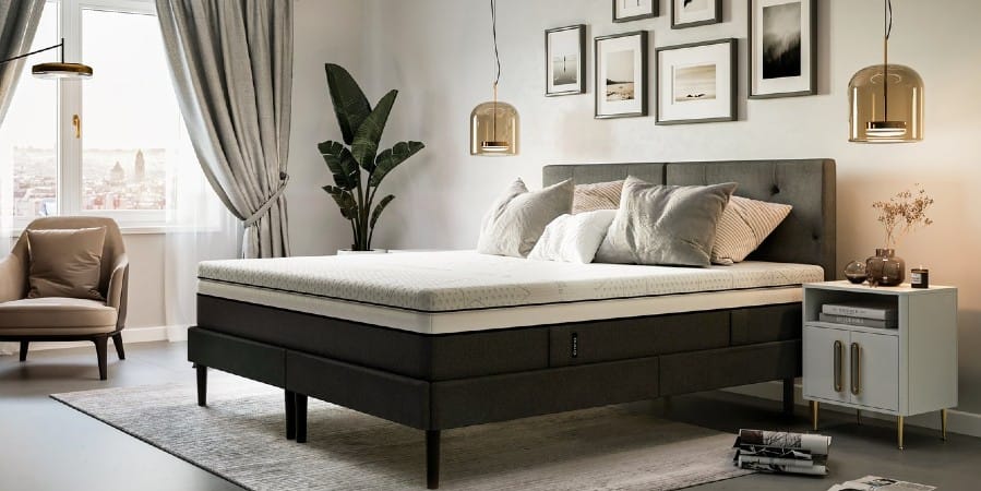 Moderne slaapkamer met groot bed, grijs hoofdeinde en witte matras en topper; twee hanglampen, nachtkastje met decoratie en plant, fauteuil bij raam met stadzicht.