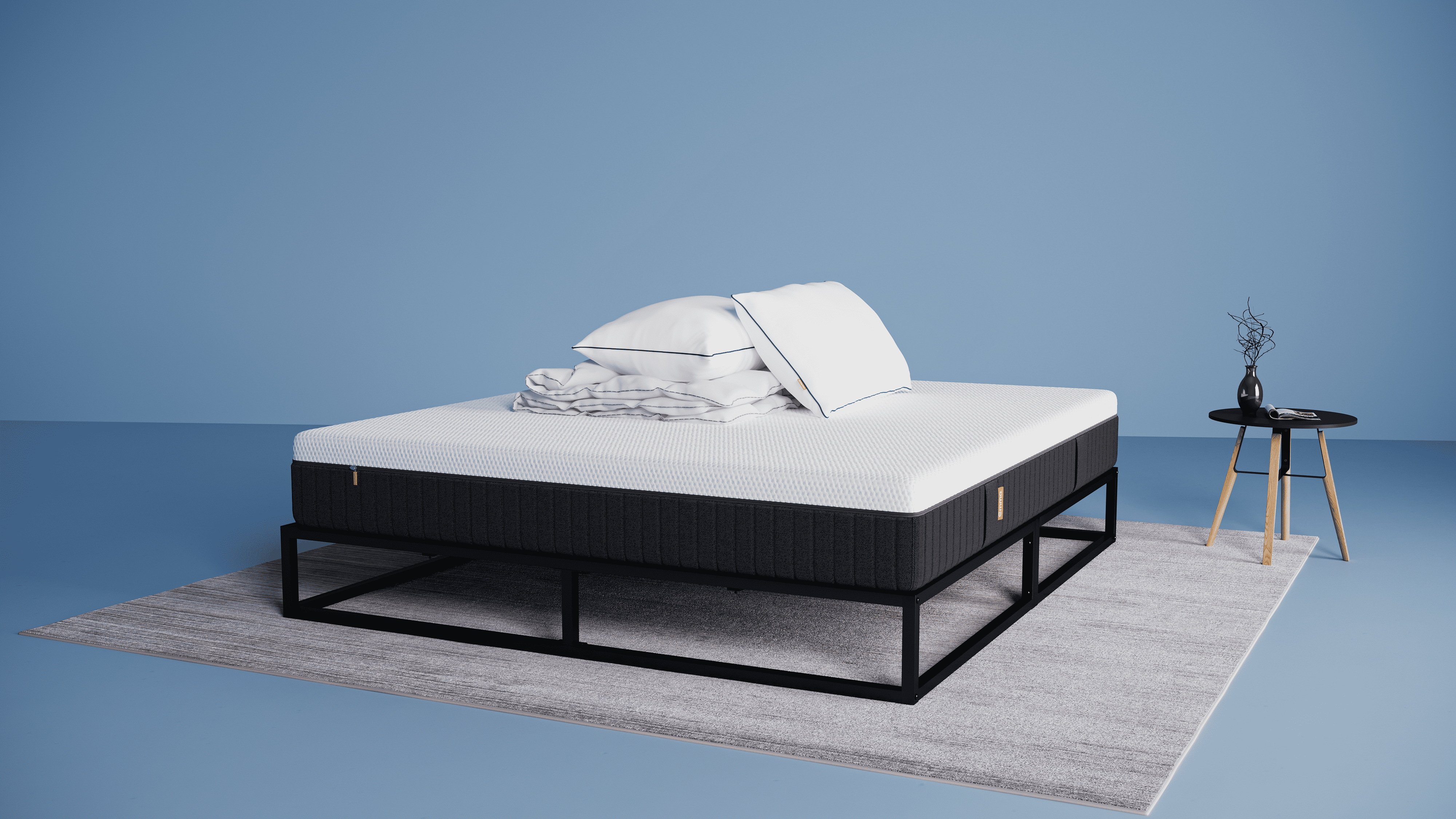 Emma Urban Deal: een modern metalen bedframe met een matras, daarop een opgevouwen wit 4-seizoenen dekbed en twee witte Emma Premium Wolkenkussens. Het bed staat op een lichtgrijs vloerkleed in een minimalistische kamer met een effen lichtblauwe achtergrond. Rechts van het bed staat een kleine ronde zwarte bijzettafel met houten poten, waarop een zwarte vaas met decoratieve takken en een tijdschrift ligt.